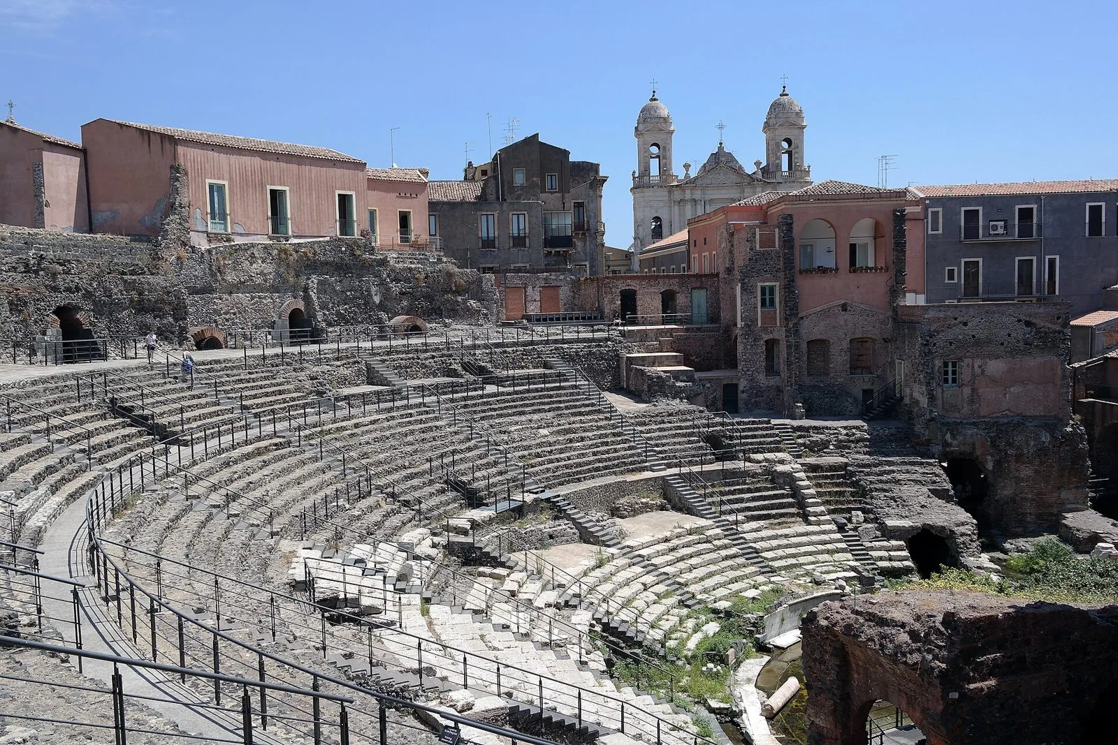 La cavea del teatro romano di Catania con le gradinate in pietra, circondata da case e dai campanili della chiesa di San Francesco d'Assisi all'Immacolata